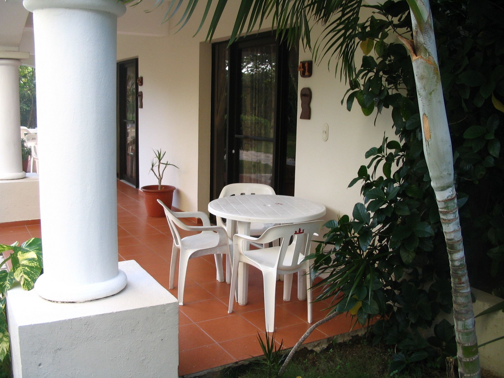 Un patio con una mesa y sillas blancas, una planta en maceta y una puerta con un cartel con número.