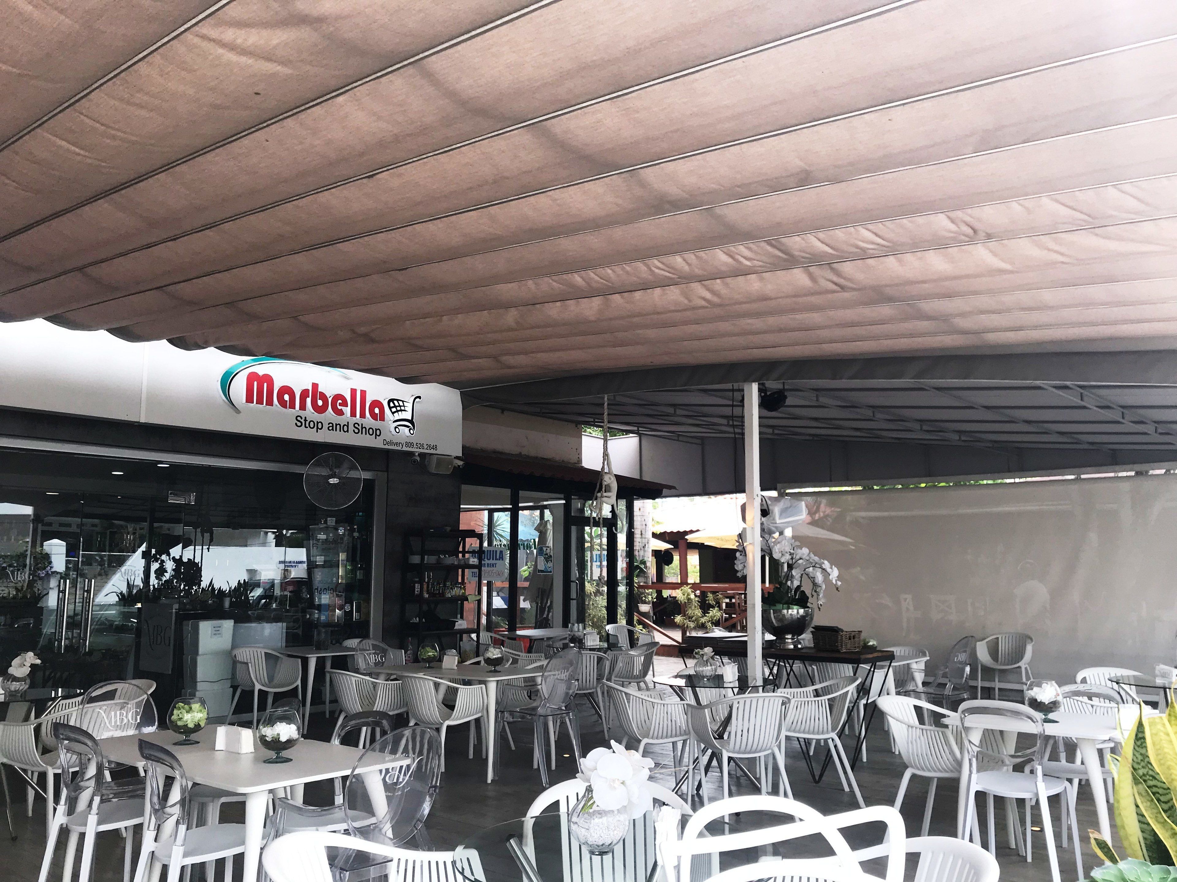 An outdoor seating area with white tables and chairs, a covered roof, and a storefront with a sign reading "Marbella Stop and Shop".