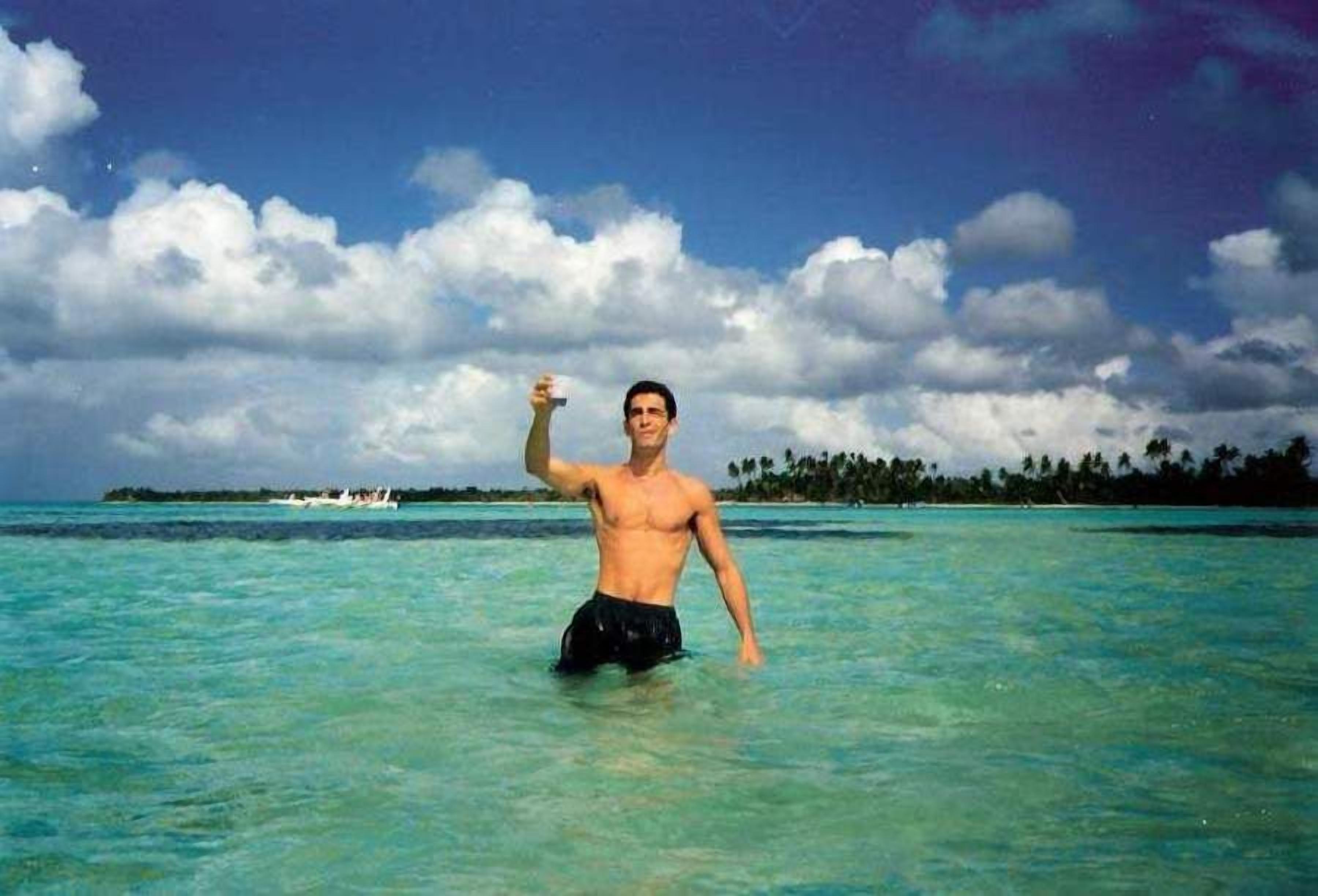 A man in a tropical setting with clear turquoise water and a distant island.