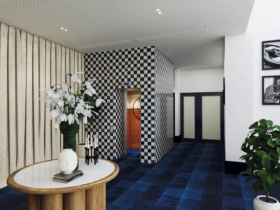 Un vestíbulo de hotel con una pared a cuadros, una mesa redonda de madera con un jarrón de flores blancas y una planta en maceta.