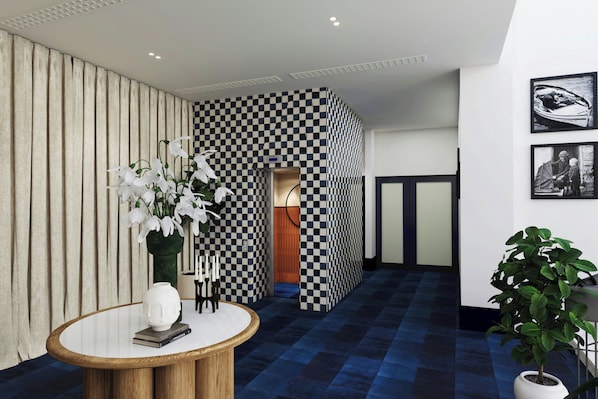 A hotel lobby with a checkered wall, a round wooden table with a vase of white flowers, and a potted plant.