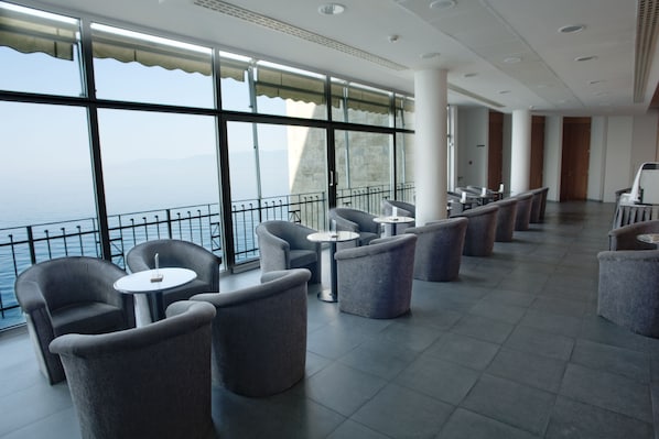 A modern indoor seating area with large windows overlooking a body of water, featuring grey upholstered chairs and small round tables. - Jadran, Rijeka, a Tribute Portfolio Hotel (Rijeka)
