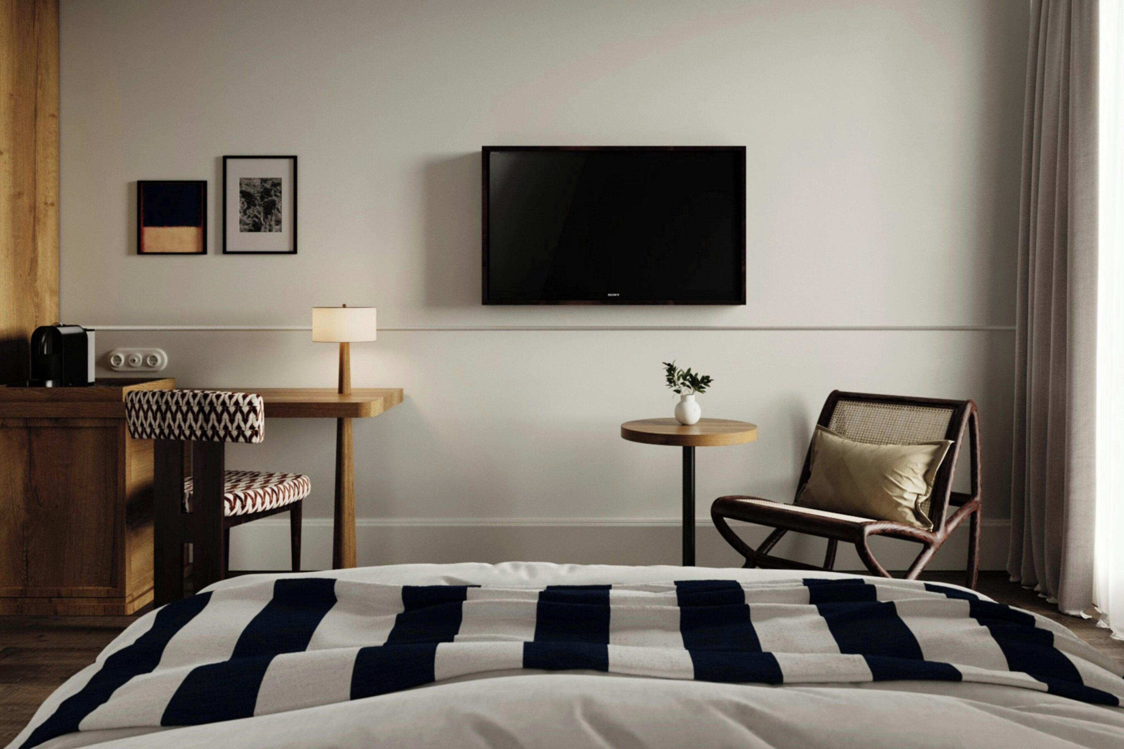 A hotel room with a bed, a desk with a lamp, a chair, a television, and two framed pictures on the wall.