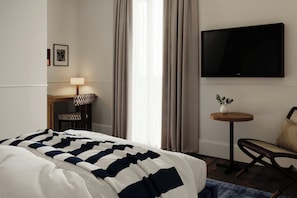 A hotel room with a bed, a TV, a desk with a lamp, a chair, and a small table with a plant.