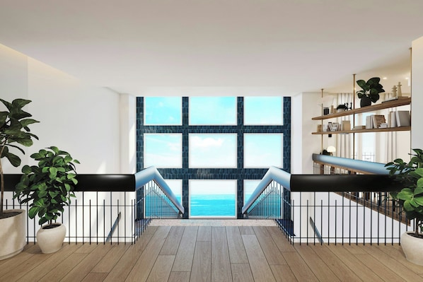 A modern interior with a view to the sea, featuring a wooden floor, black railing, and a bookshelf.