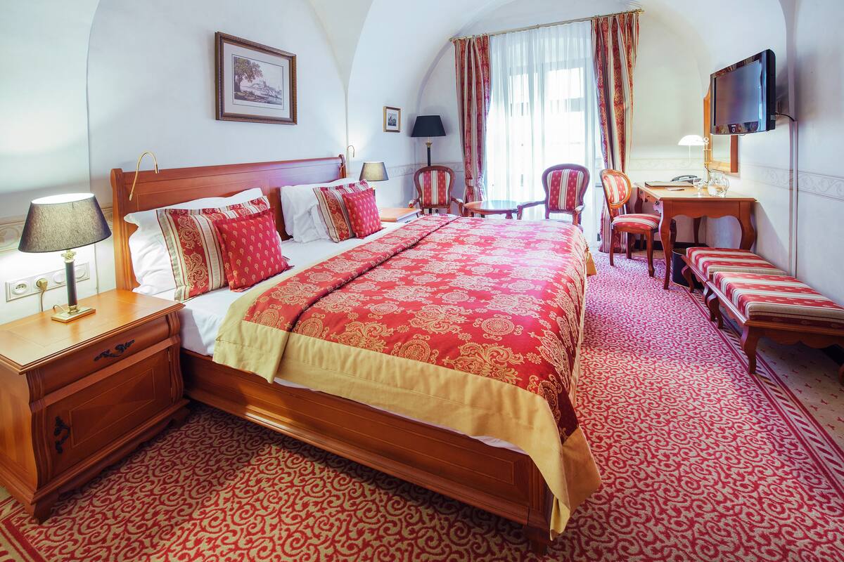 Ein Hotelzimmer mit einem großen Bett, einem Schreibtisch mit Stuhl, einem Fernseher und einem gemusterten Teppich.