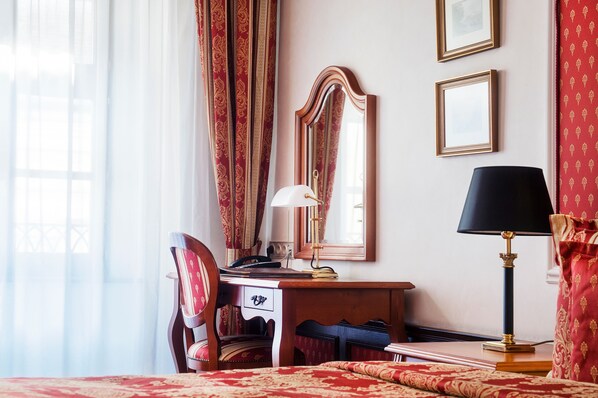 A hotel room with a wooden desk, a chair, a mirror, a lamp, and a bed with a red patterned bedspread.