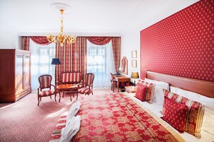 Habitación de hotel con una cama grande, un armario de madera, un escritorio con silla, una araña y un ventanal con cortinas.