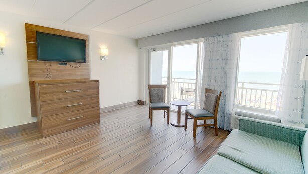 A hotel room with a wooden floor, a sofa, a TV mounted on a wooden panel, two chairs, and a balcony with a view. - Marjac Suites Virginia Beach Oceanfront (Virginia Beach)