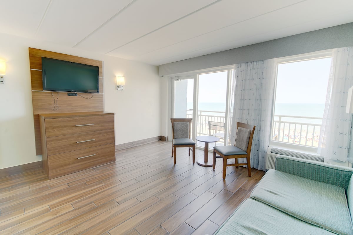 Habitación de hotel con piso de madera, un sofá, un televisor montado en un panel de madera, dos sillas y un balcón con vistas.