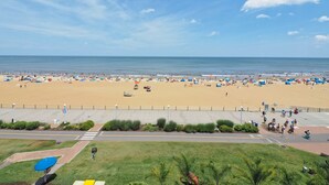 A beach with a paved walkway and a grassy area with a blue umbrella. - Marjac Suites Virginia Beach Oceanfront (Virginia Beach)