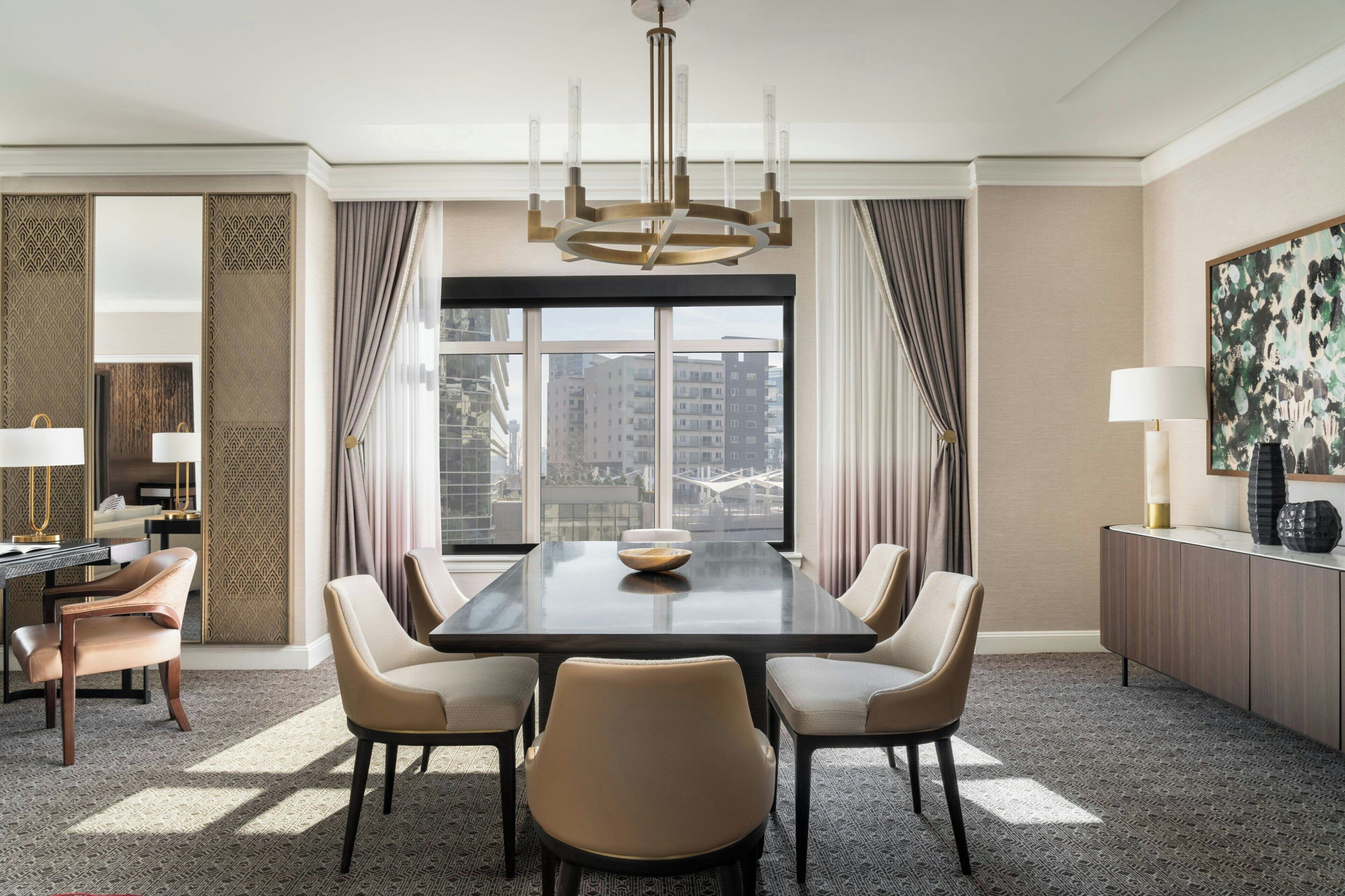 Quarto de hotel com uma grande janela, mesa de jantar com cadeiras, lustre e vista para edifícios.