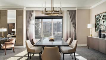 Quarto de hotel com uma grande janela, mesa de jantar com cadeiras, lustre e vista para edifícios.