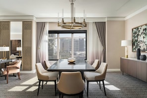 A hotel room with a large window, a dining table with chairs, a chandelier, and a view of buildings.
