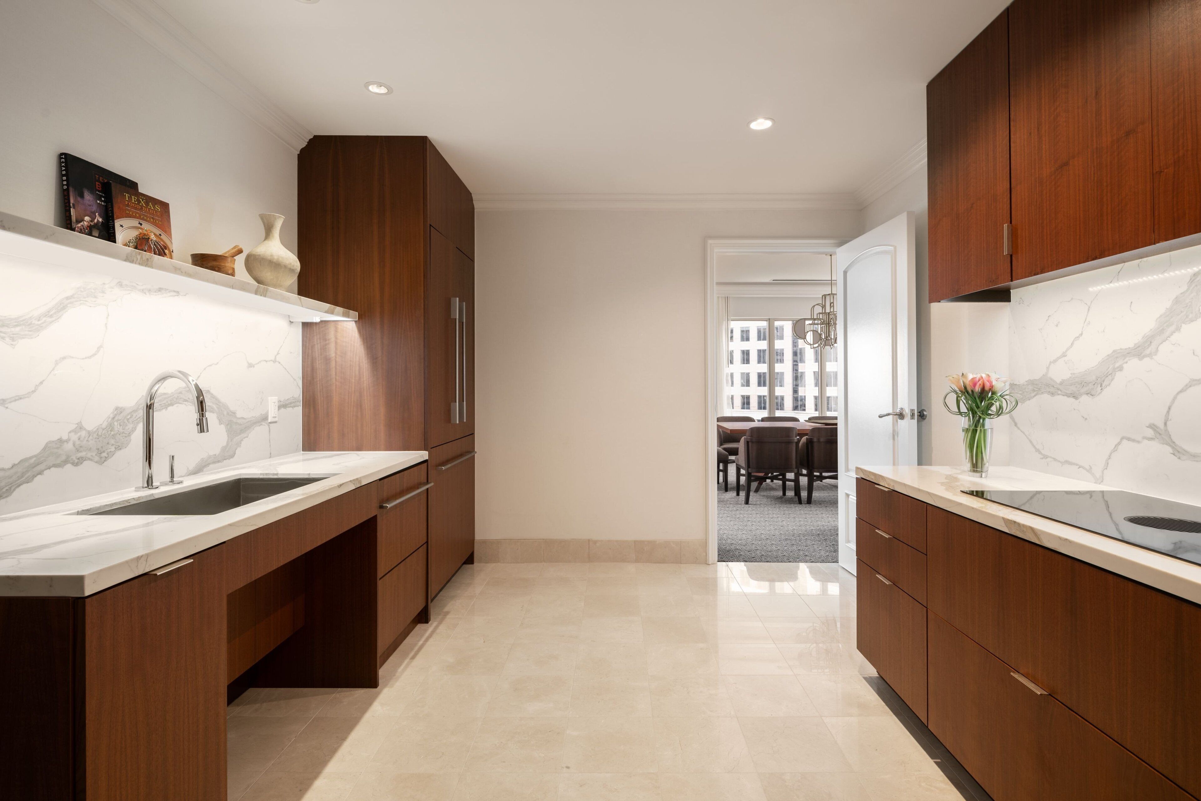 A modern kitchen with marble countertops, wooden cabinets, and a view into the dining area.