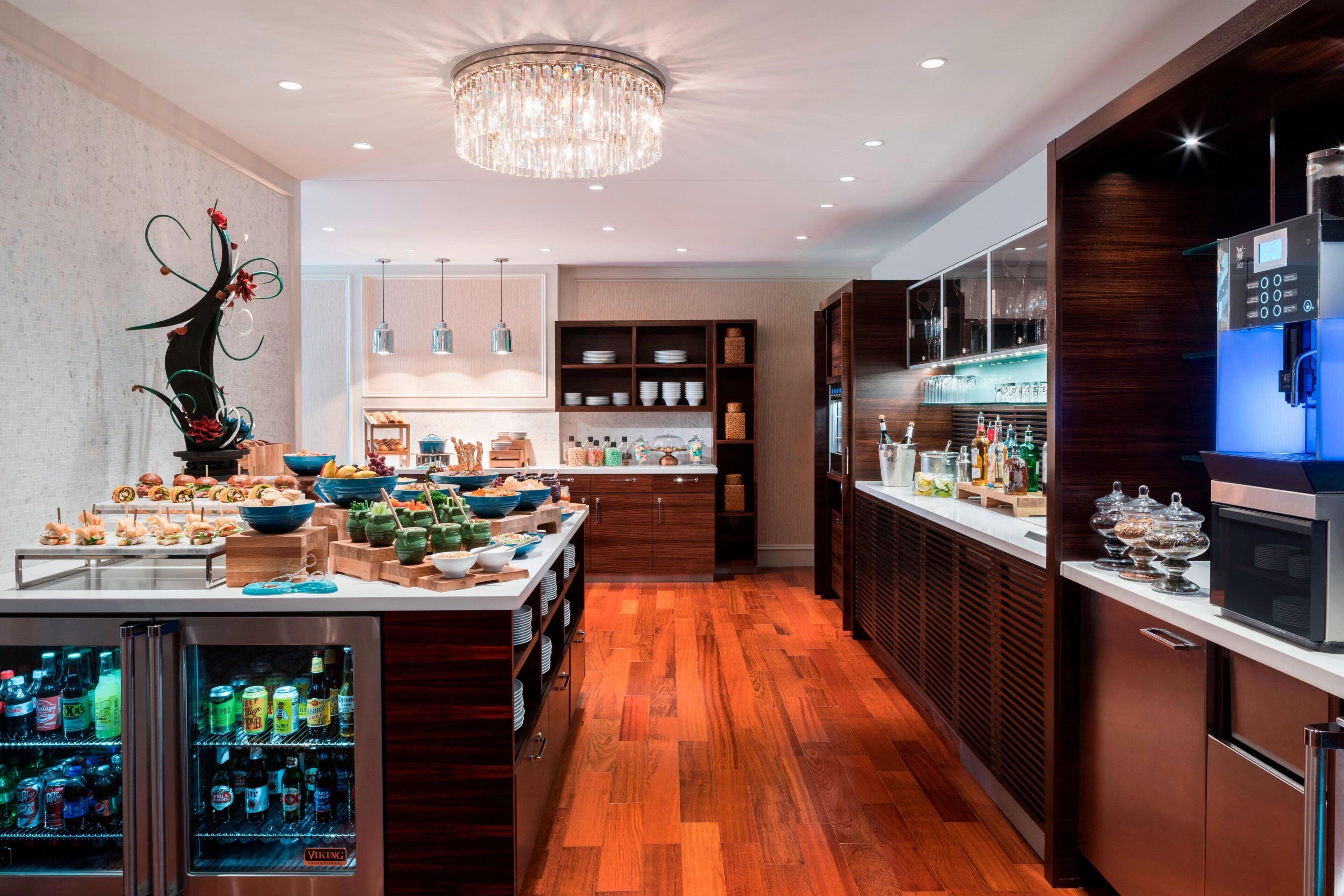 A modern kitchen with a wooden floor, a central island with food and drinks, a refrigerator, and a microwave.