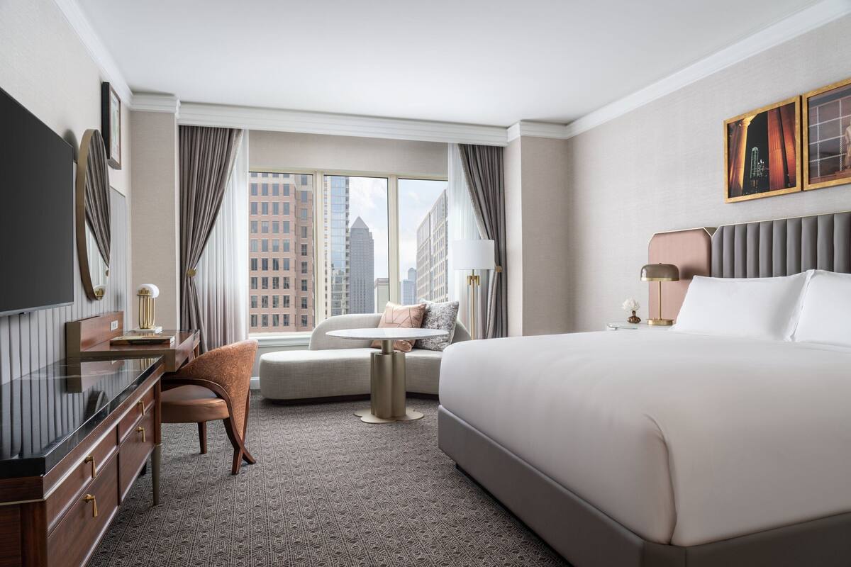 A hotel room with a large bed, a desk with a flat-screen TV, a sofa, and a view of city buildings.
