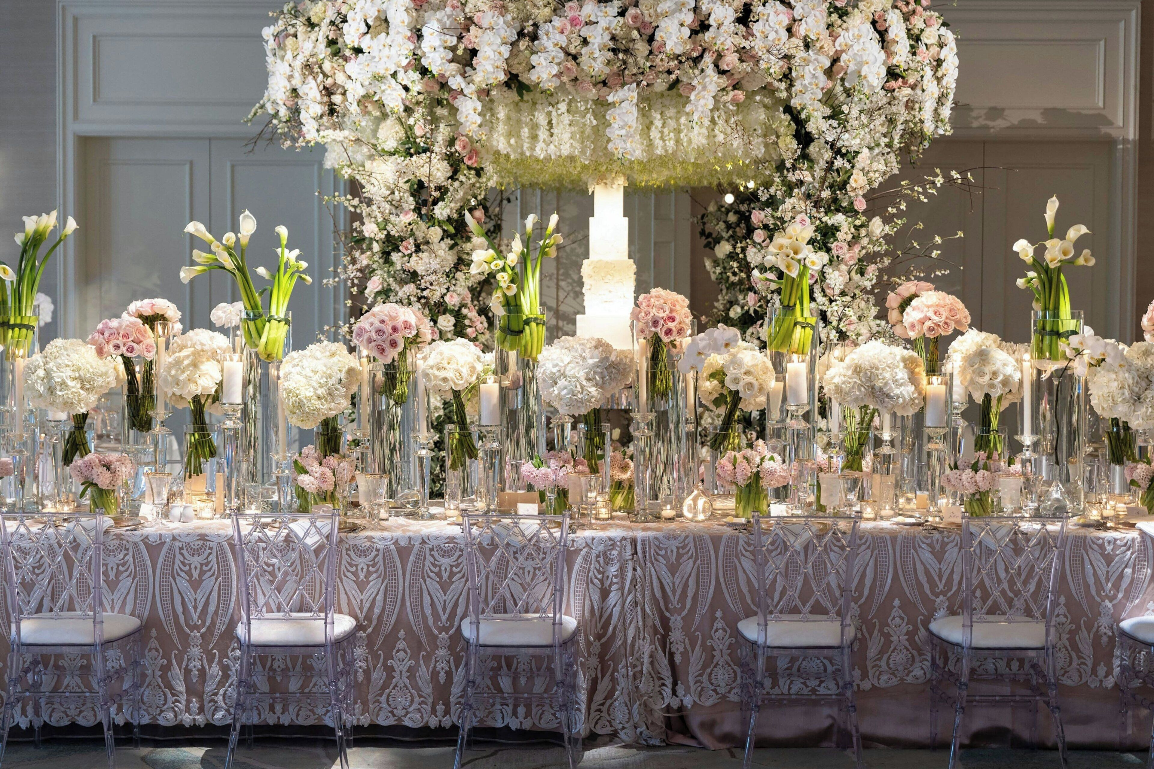 A long table with clear chairs, adorned with floral arrangements and candles, set against a wall with a decorative arch.
