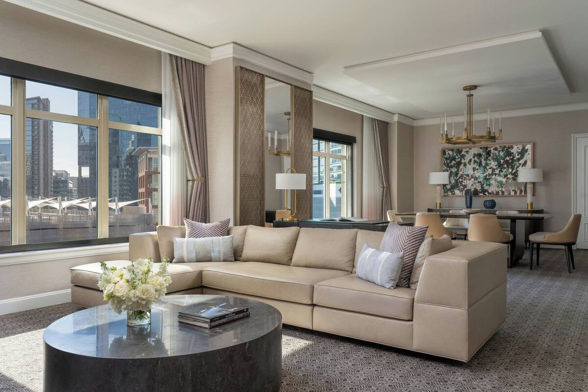 A modern living room with a beige sectional sofa, a round coffee table, and a dining area with a chandelier.