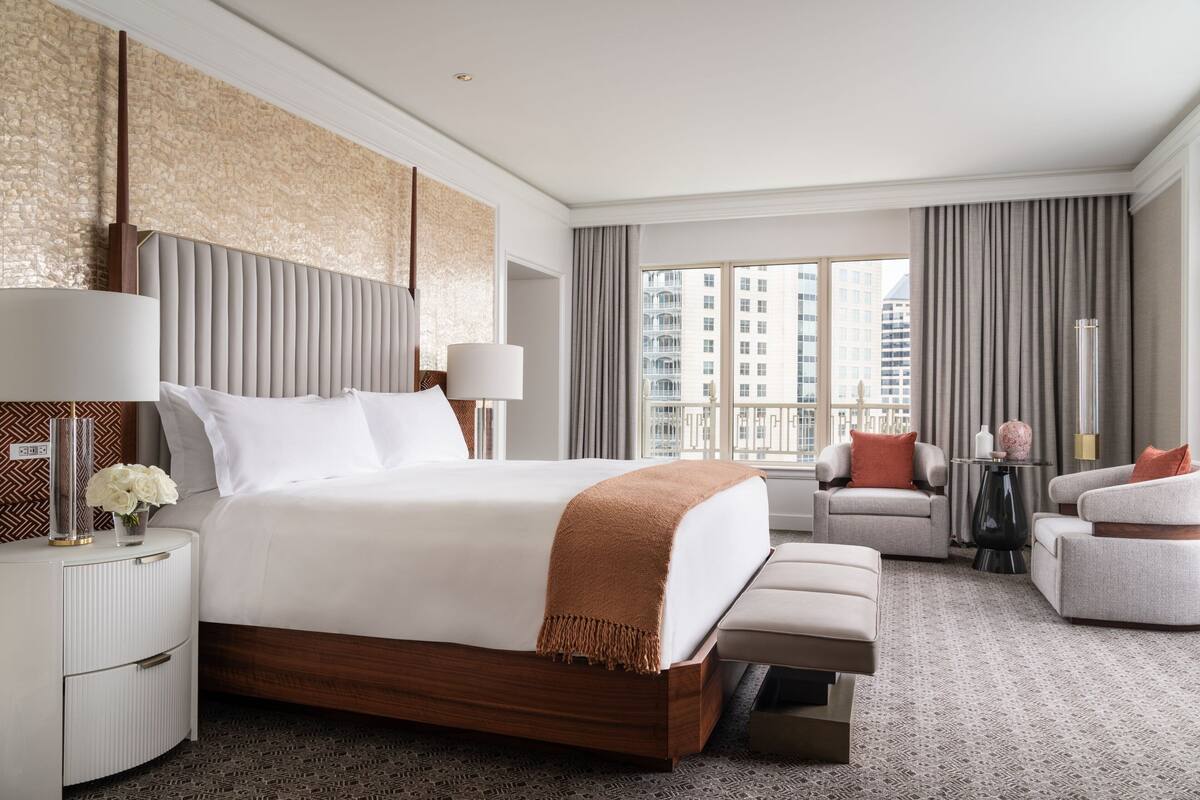 A modern hotel room with a large bed, a seating area, and a view of the cityscape.