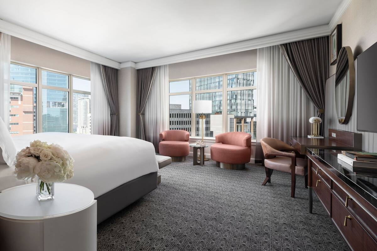 A hotel room with a large bed, two pink armchairs, a small table, a TV, and a view of the city through large windows.