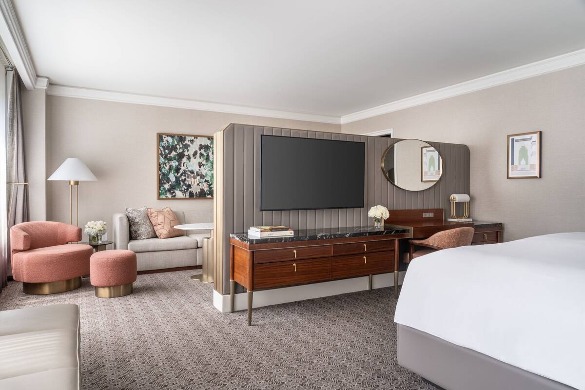 A modern hotel room with a large bed, a sofa, a desk with a flat-screen TV, and a vanity with a mirror.