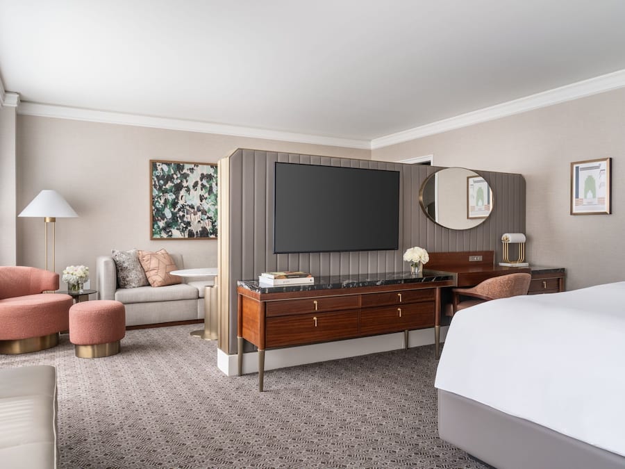 A modern hotel room with a large bed, a sofa, a desk with a flat-screen TV, and a vanity with a mirror.