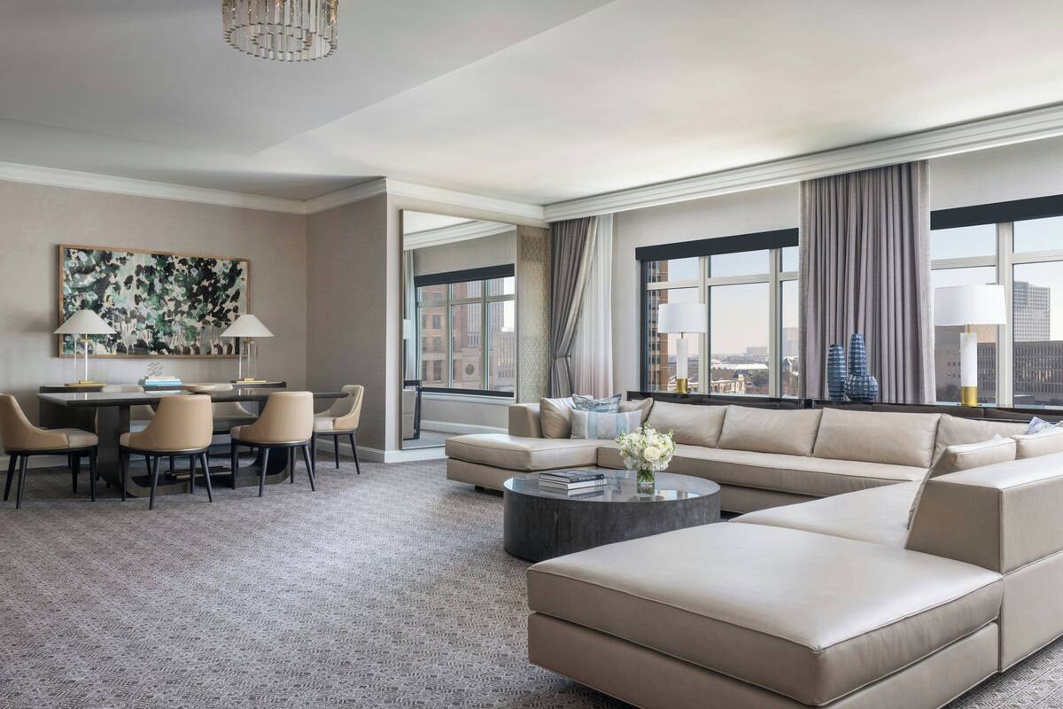 A modern hotel room with a large sectional sofa, a dining area with chairs, a coffee table, and a city view through large windows.