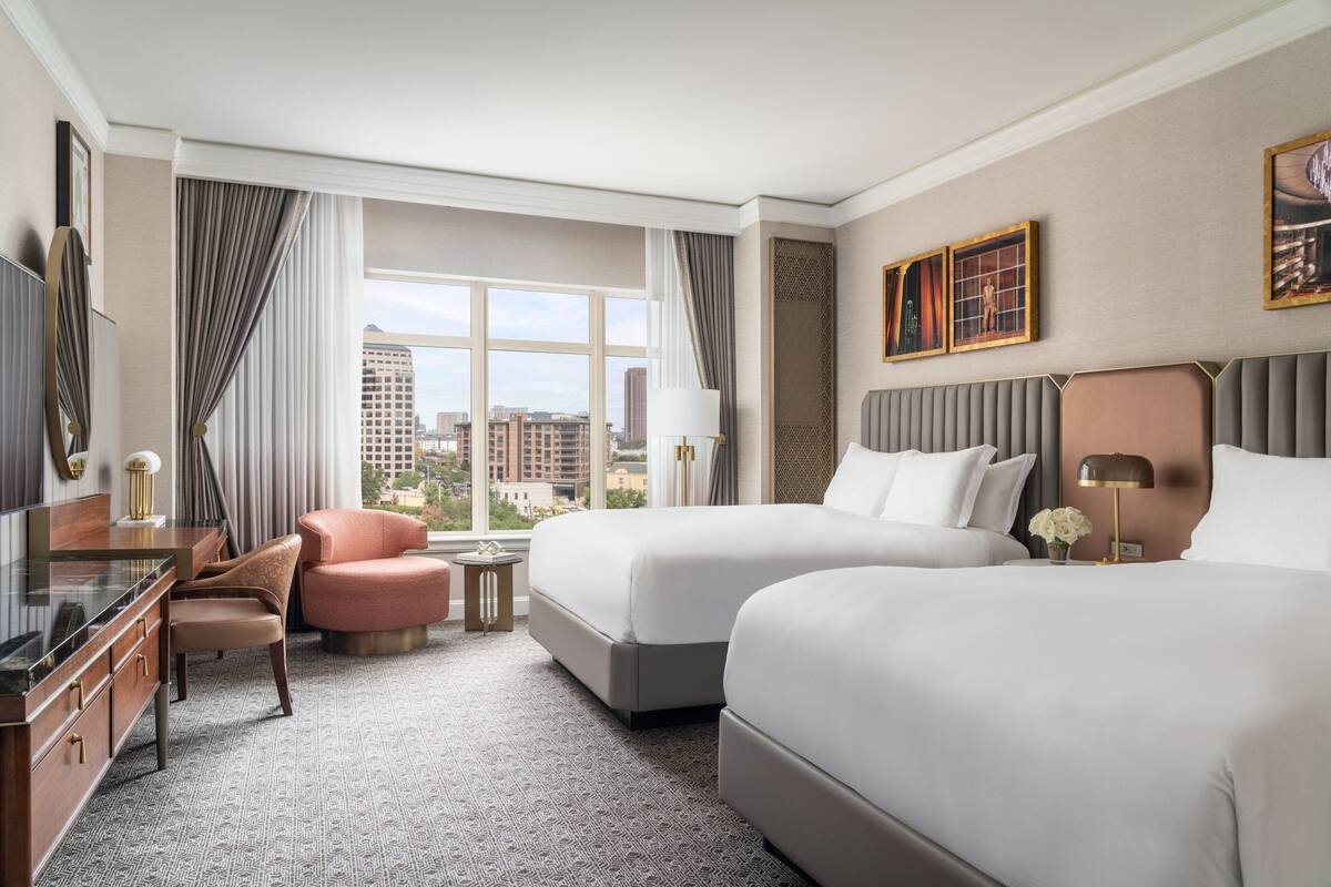 A hotel room with two beds, a desk, a chair, a TV, and a large window with curtains.