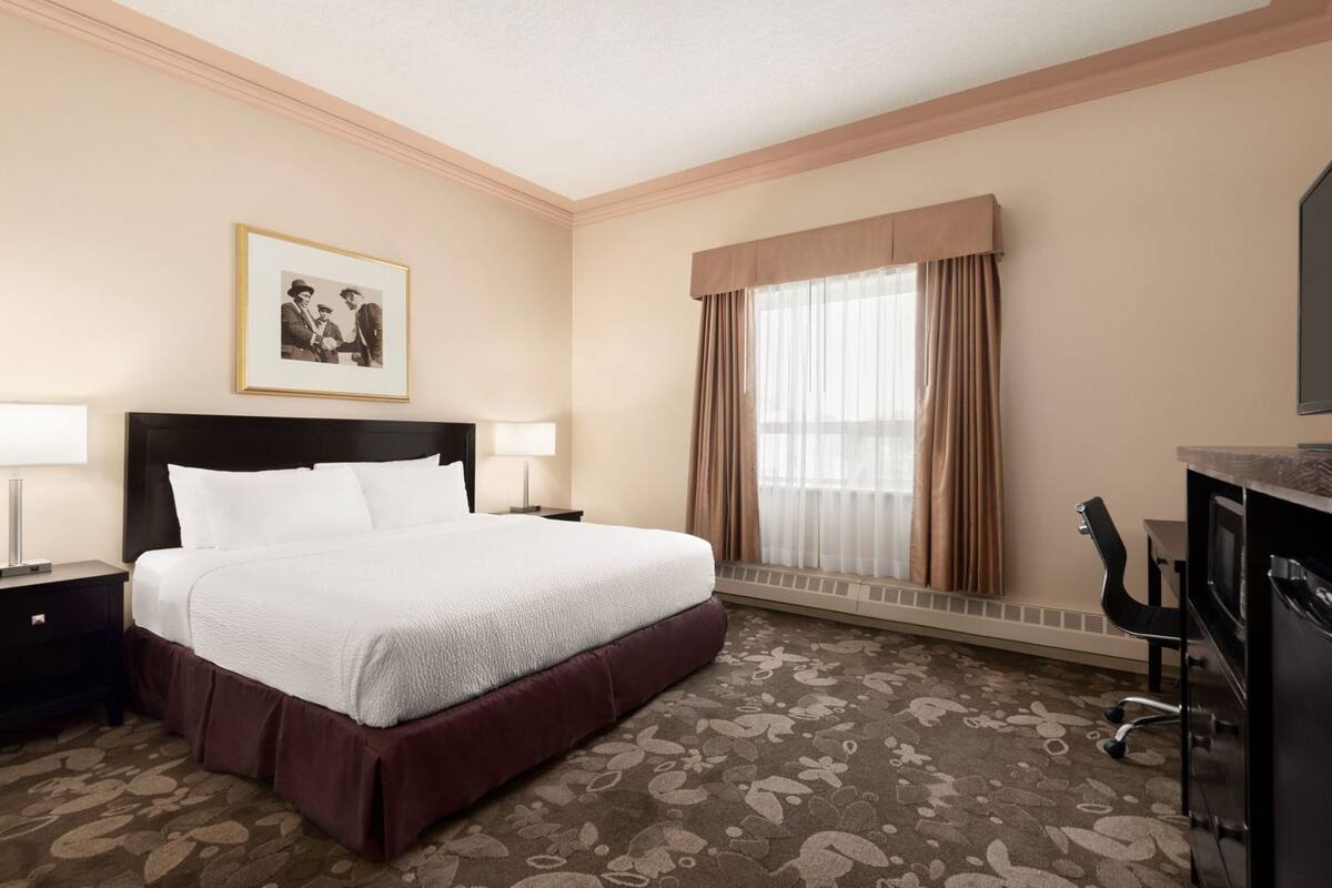 Una habitación de hotel con una cama grande, dos mesitas de noche con lámparas, un escritorio con una silla, una ventana con cortinas y un cuadro en la pared.
