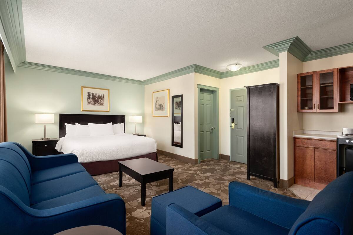 Habitación de hotel con una cama, dos sillones azules, una mesita, un escritorio, un armario y una kitchenette.