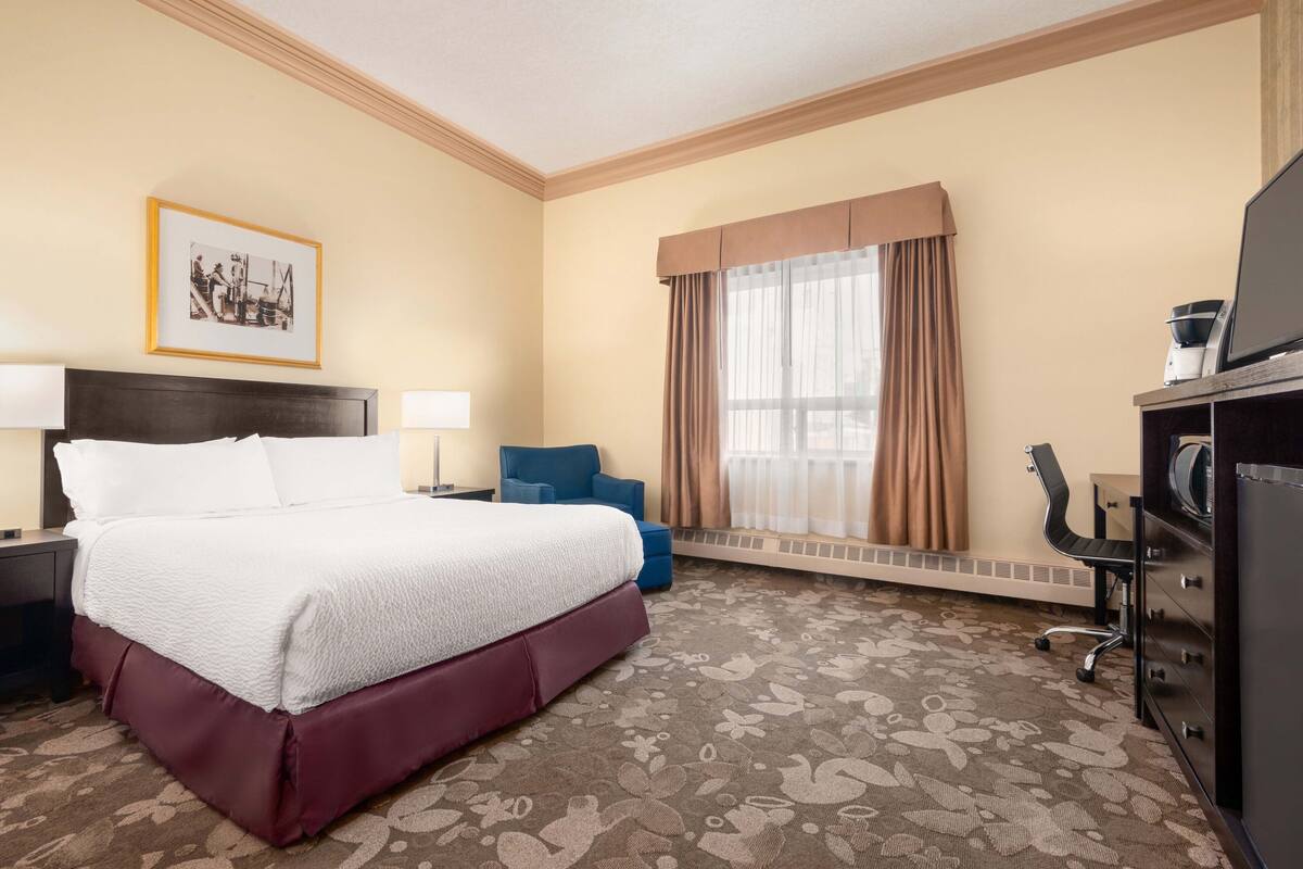 Habitación de hotel con una cama grande, un escritorio con computadora, una silla, un ventanal con cortinas y un radiador.