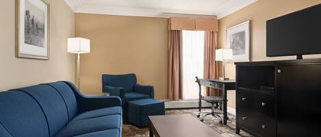 A hotel room with a blue sofa, a small table, a desk with a chair, a television, and a filing cabinet.