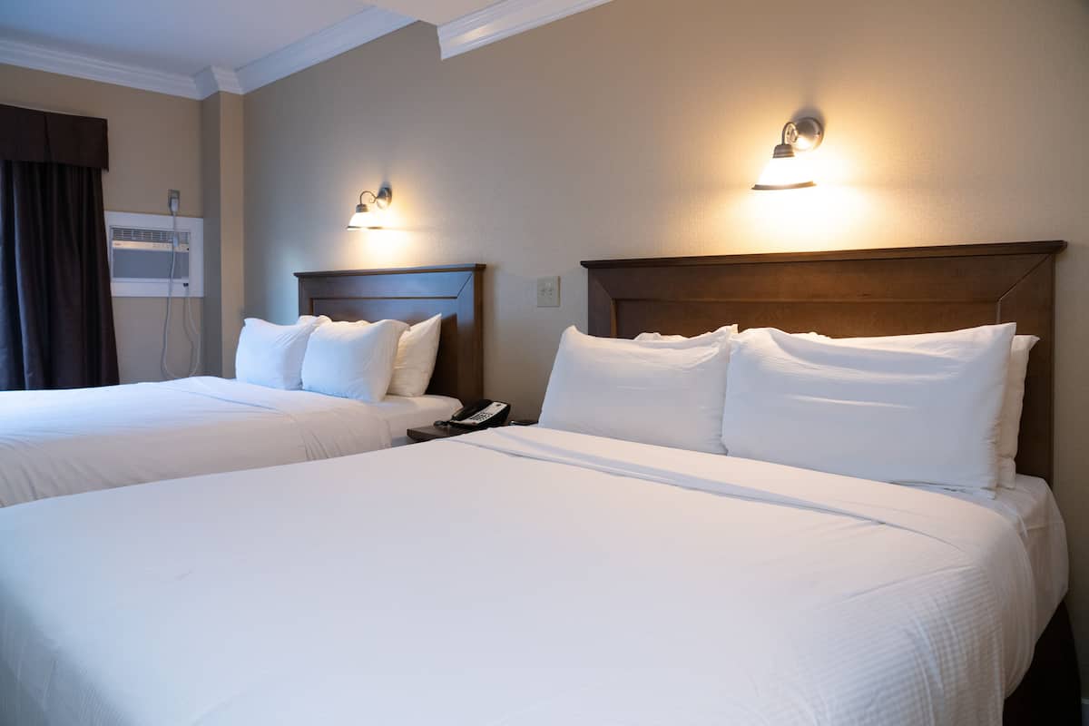 Habitación de hotel con dos camas, cada una con sábanas blancas y cabeceros. Se observa un aire acondicionado de pared y un teléfono en la mesita de noche.