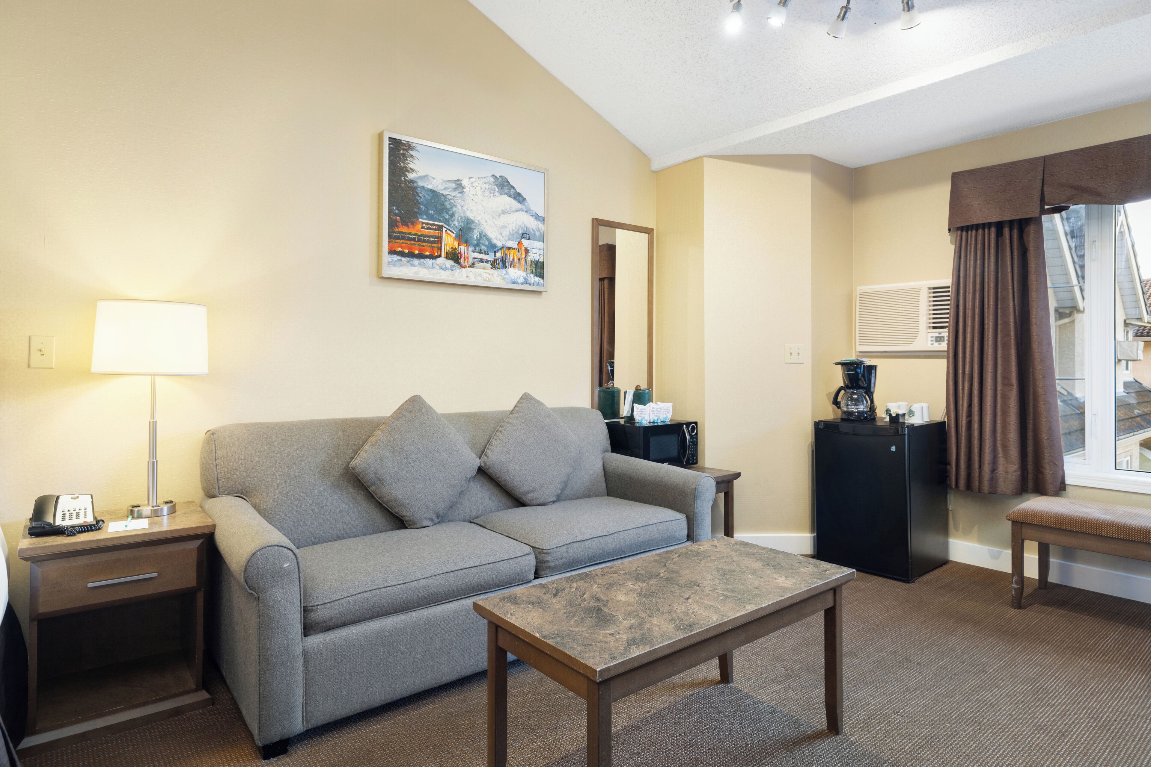 Une chambre d’hôtel avec un canapé, une table basse, un bureau avec une lampe, un tableau au mur et une vue sur l’extérieur.