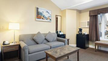 A hotel room with a sofa, a coffee table, a desk with a lamp, a painting on the wall, and a view of the outside.