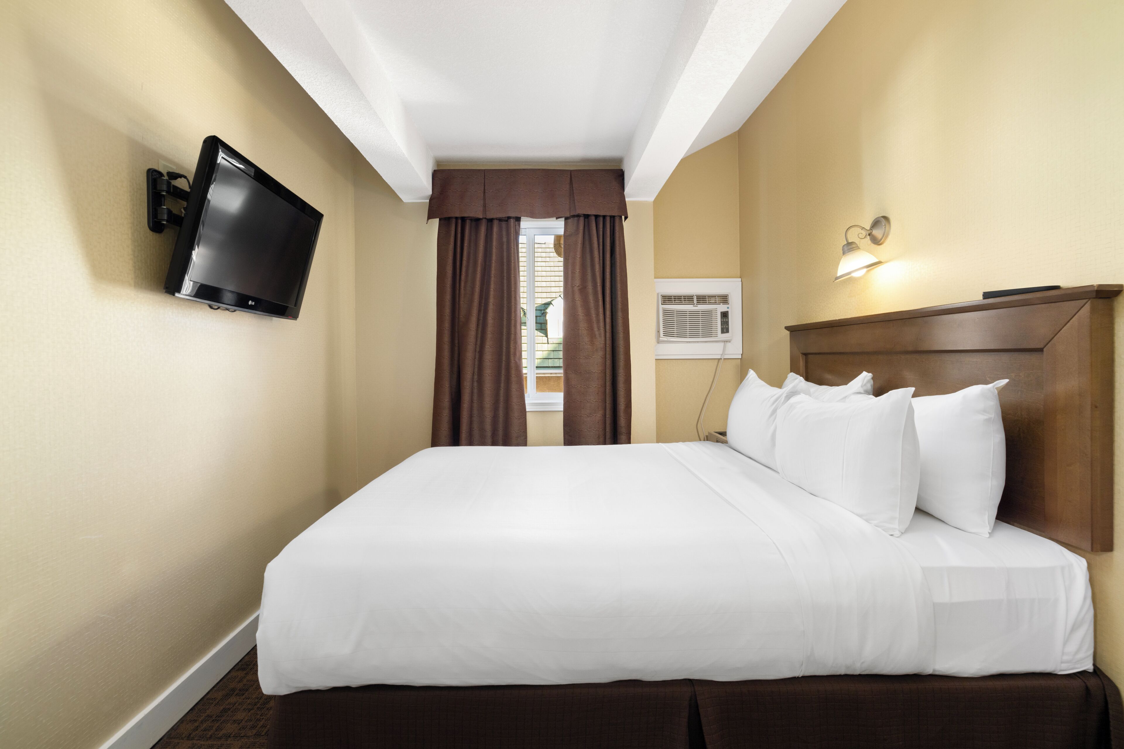 A hotel room with a large bed, a flat-screen TV mounted on the wall, a ceiling fan, and a window with curtains.