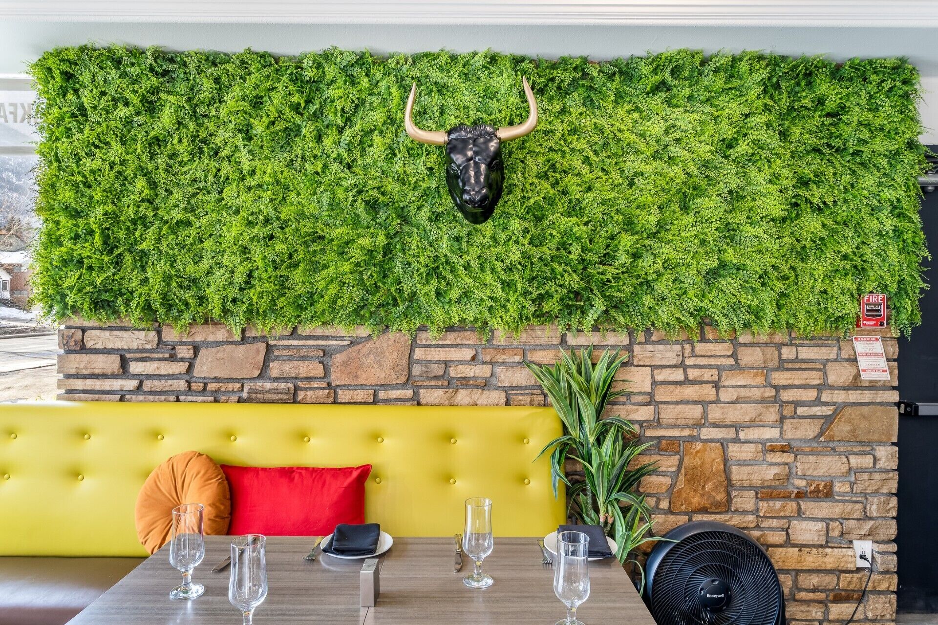 Uma área de refeições com parede de pedra, parede verde decorada com cabeça de touro, sofá amarelo e mesa posta com copos e talheres.