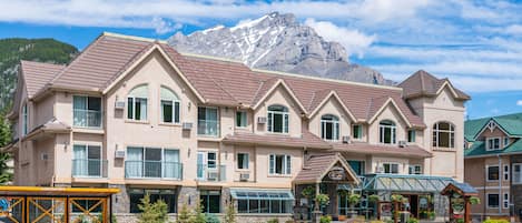 A multi-story hotel with a mountain backdrop, featuring a central entrance with a glass canopy, multiple windows, and a stone facade.
