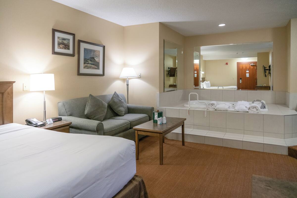 Habitación de hotel con cama, sofá, mesita, lámpara, espejo y baño con bañera y toallas.