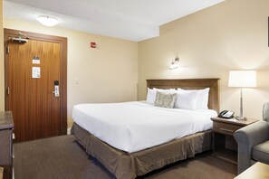 A hotel room with a wooden door, a bed with white bedding, a nightstand with a lamp, and a chair. - Irwin's Mountain Inn (Banff)