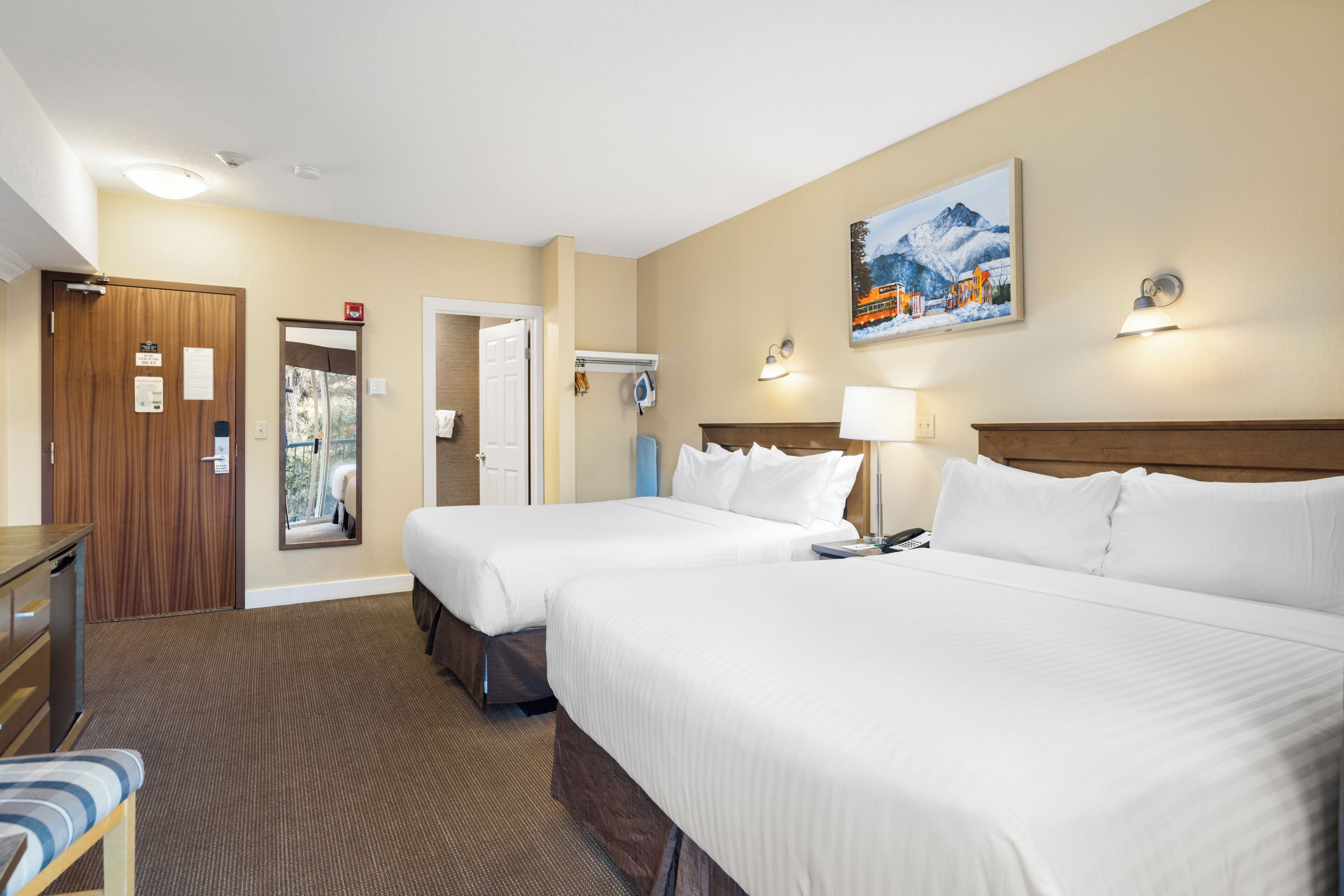 Une chambre d’hôtel avec deux lits, un bureau, une chaise, un miroir, un tableau et une lampe fixée au mur.