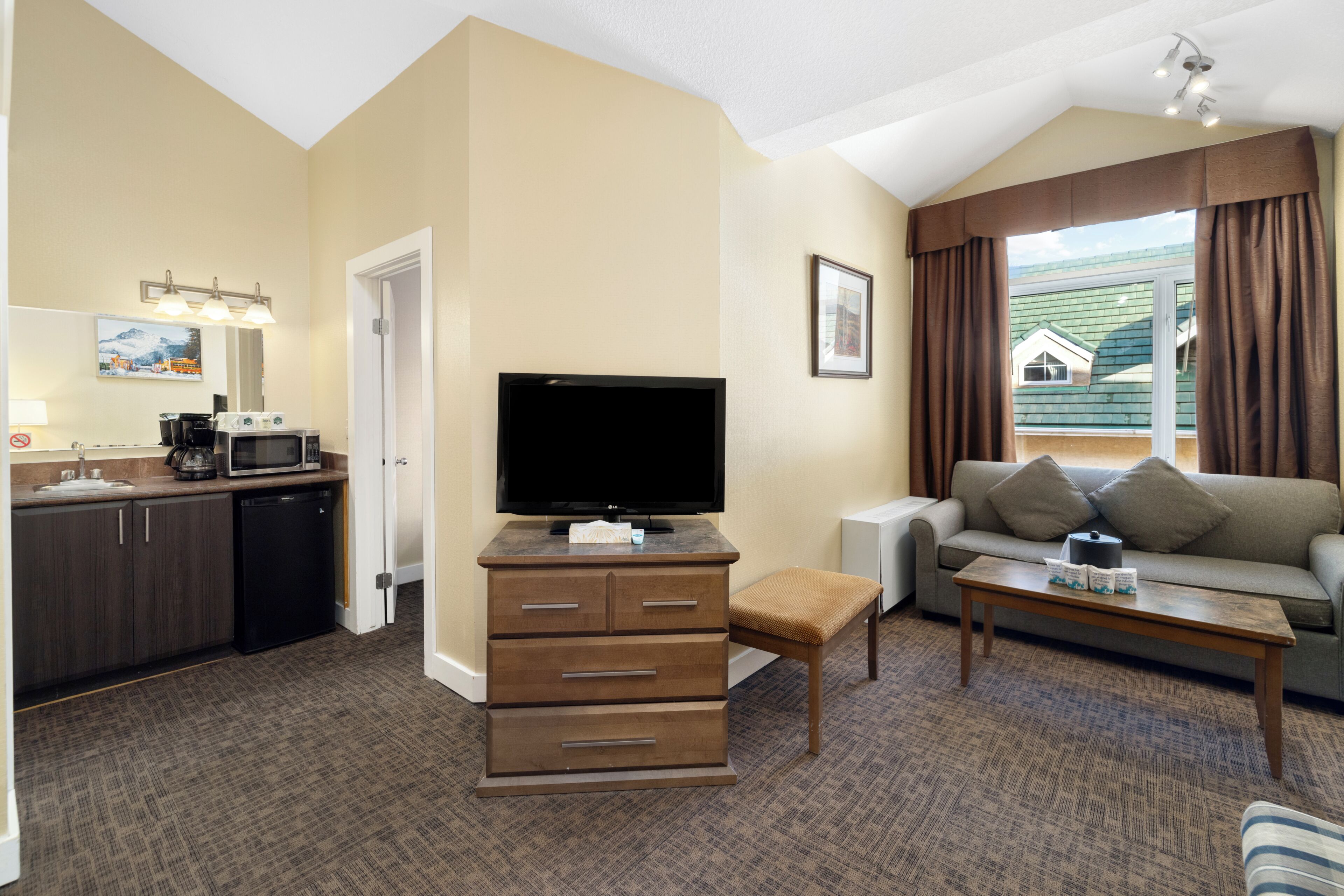 Quarto de hotel com cozinha compacta, televisão de tela plana, sofá, mesa de centro e janela com cortinas.
