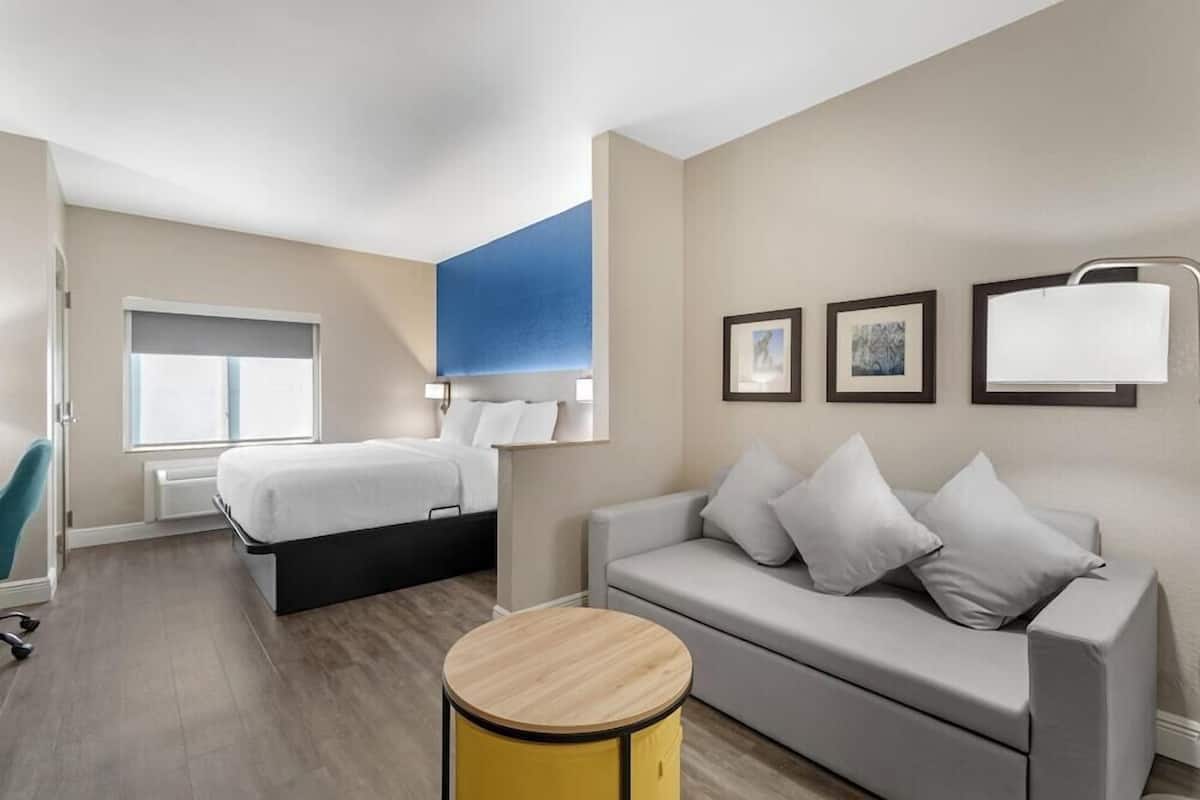 Una habitación de hotel moderna con cama, sofá, mesa redonda de madera y ventana con persianas.