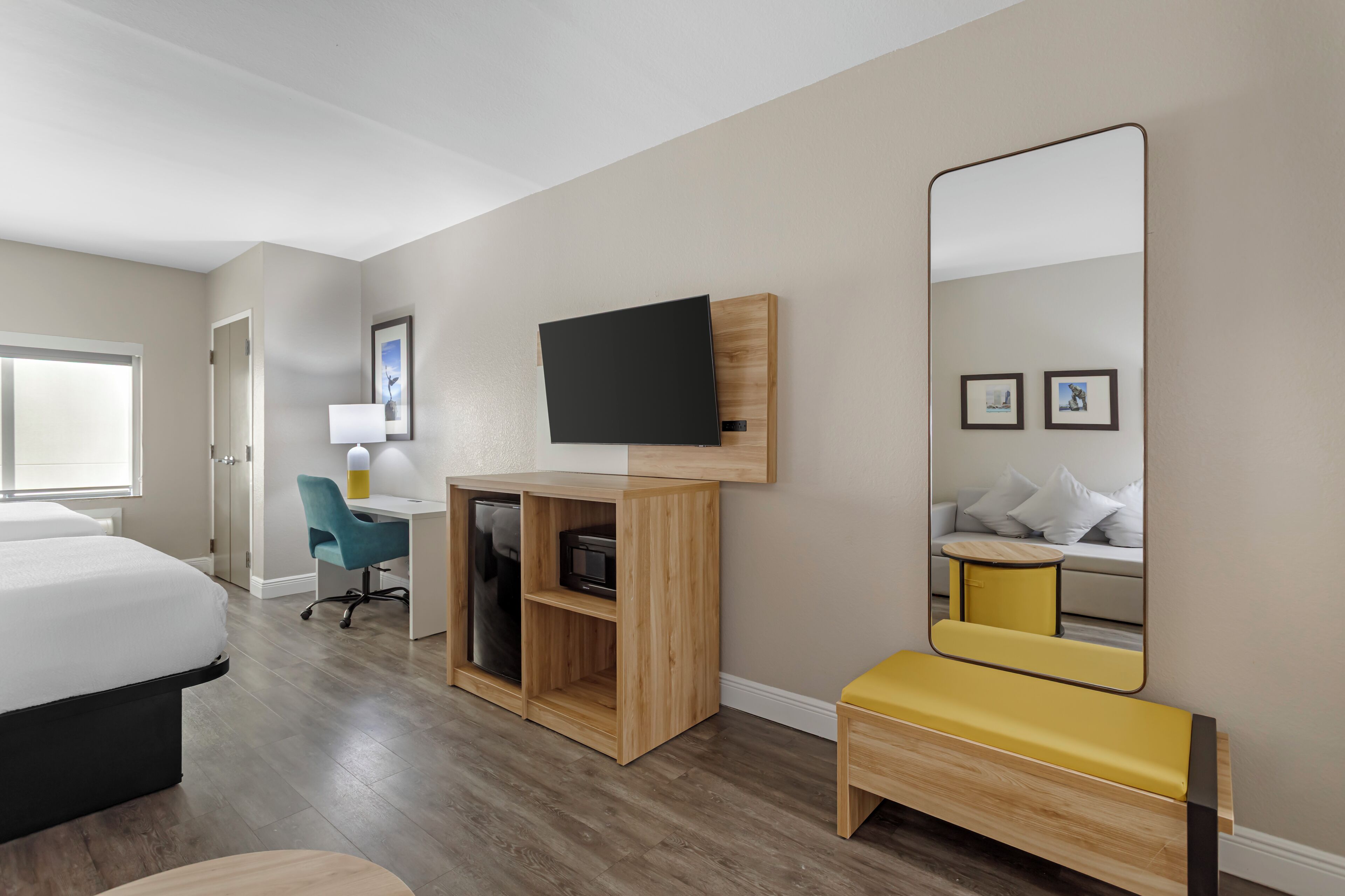 Quarto de hotel com cama, escrivaninha com cadeira, televisão, espelho e um banco.