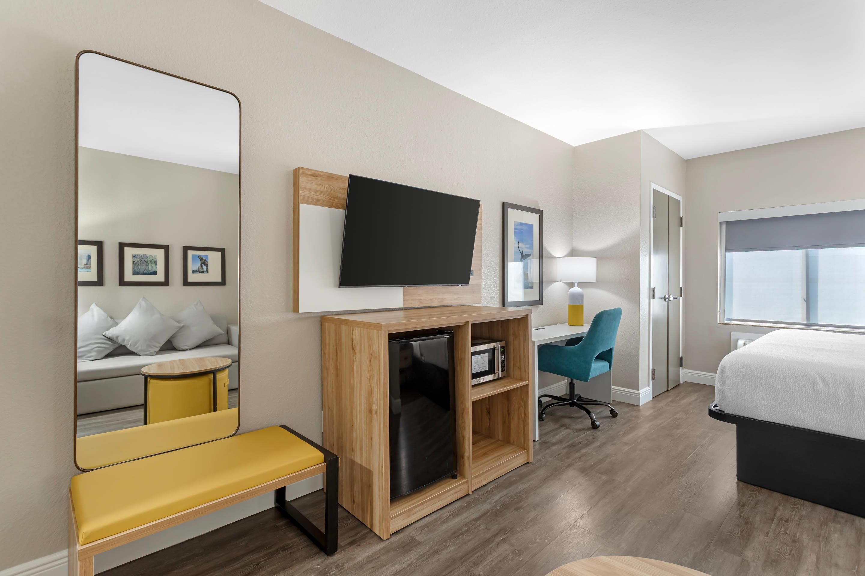 Quarto de hotel com cama, escrivaninha com cadeira, televisão, espelho, banco e micro-ondas.