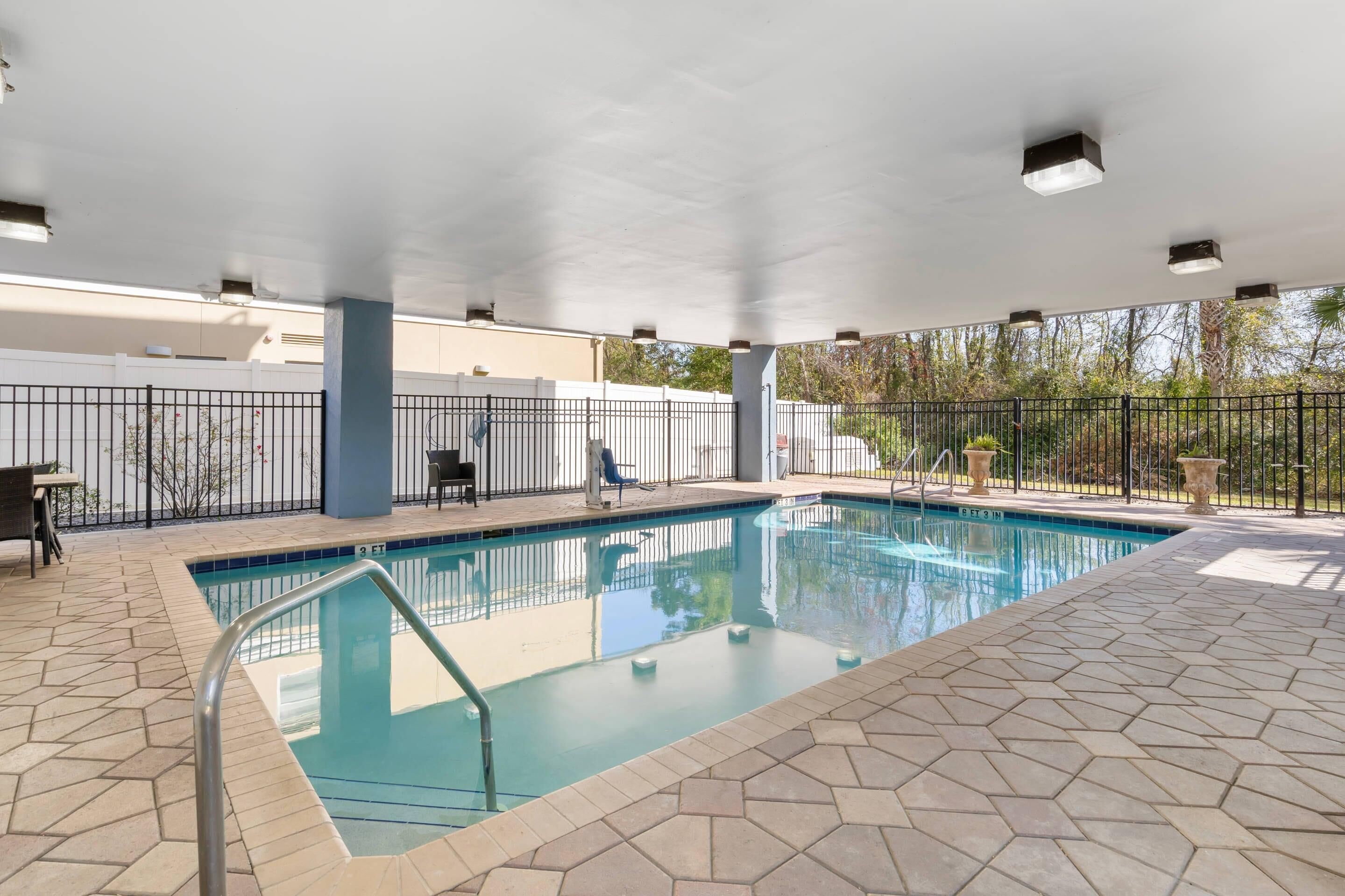 An indoor swimming pool with a tiled floor, a metal pool ladder, and a covered area with a white ceiling and ceiling lights.