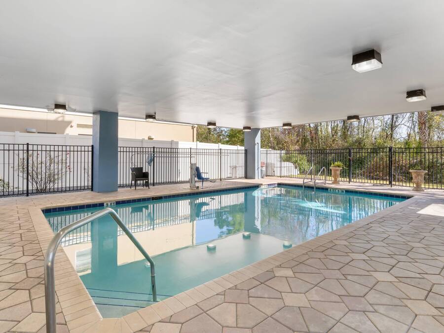 An indoor swimming pool with a tiled floor, a metal pool ladder, and a covered area with a white ceiling and ceiling lights.