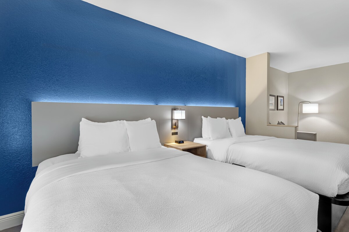 Habitación de hotel con dos camas, una mesita de noche con lámpara y un espejo en la pared.
