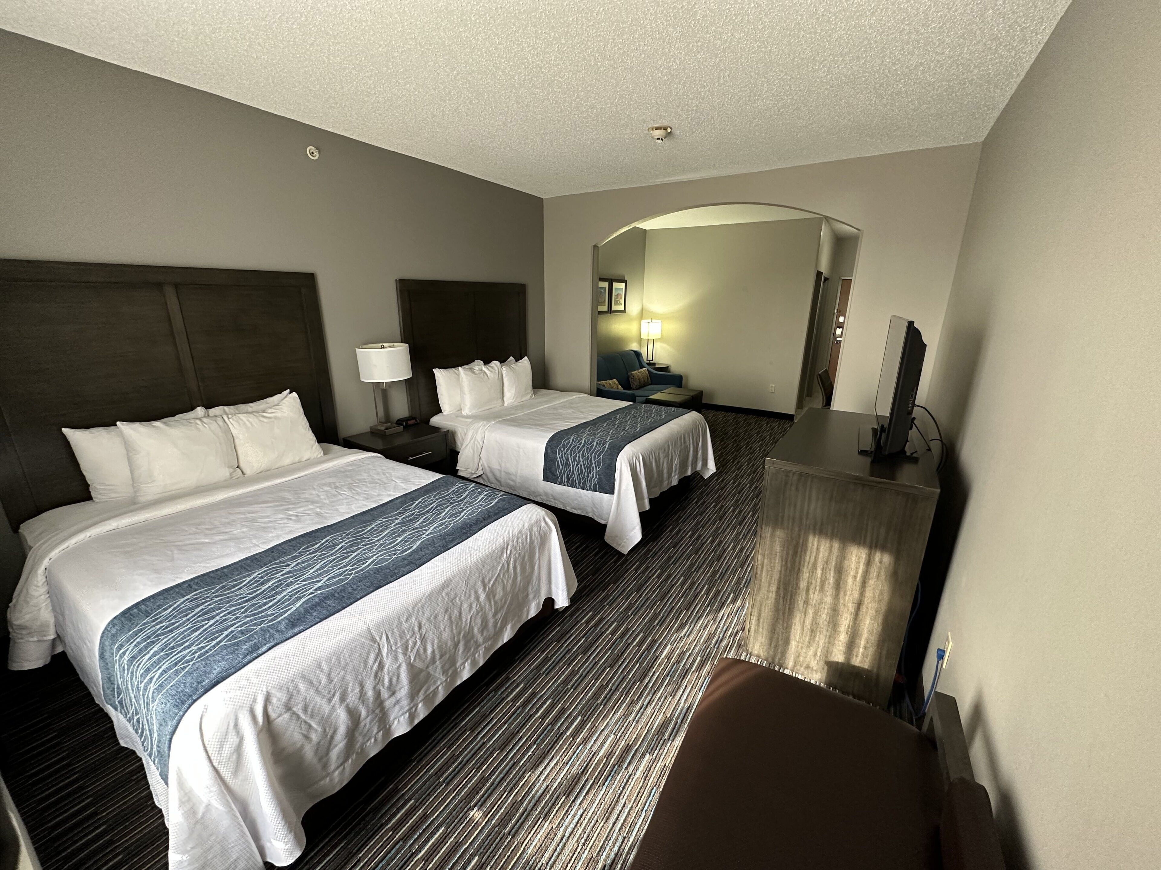A hotel room with two beds, a desk, a chair, a lamp, and a TV.
